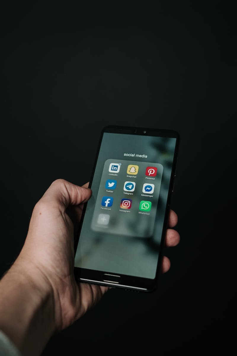 Close-up of a hand holding a smartphone displaying various social media app icons on a dark background.