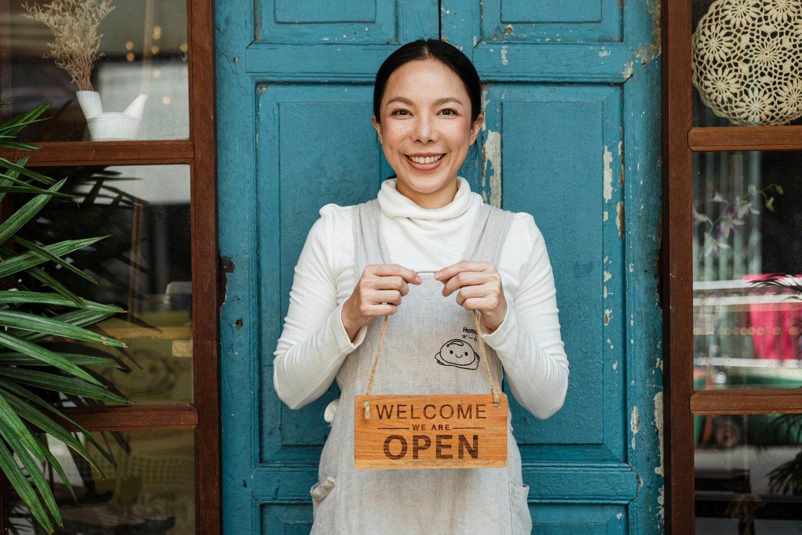 Cheerful ethnic female cafeteria owner in apron demonstrating cardboard signboard while standing near blue shabby door and windows after starting own business and looking at camera